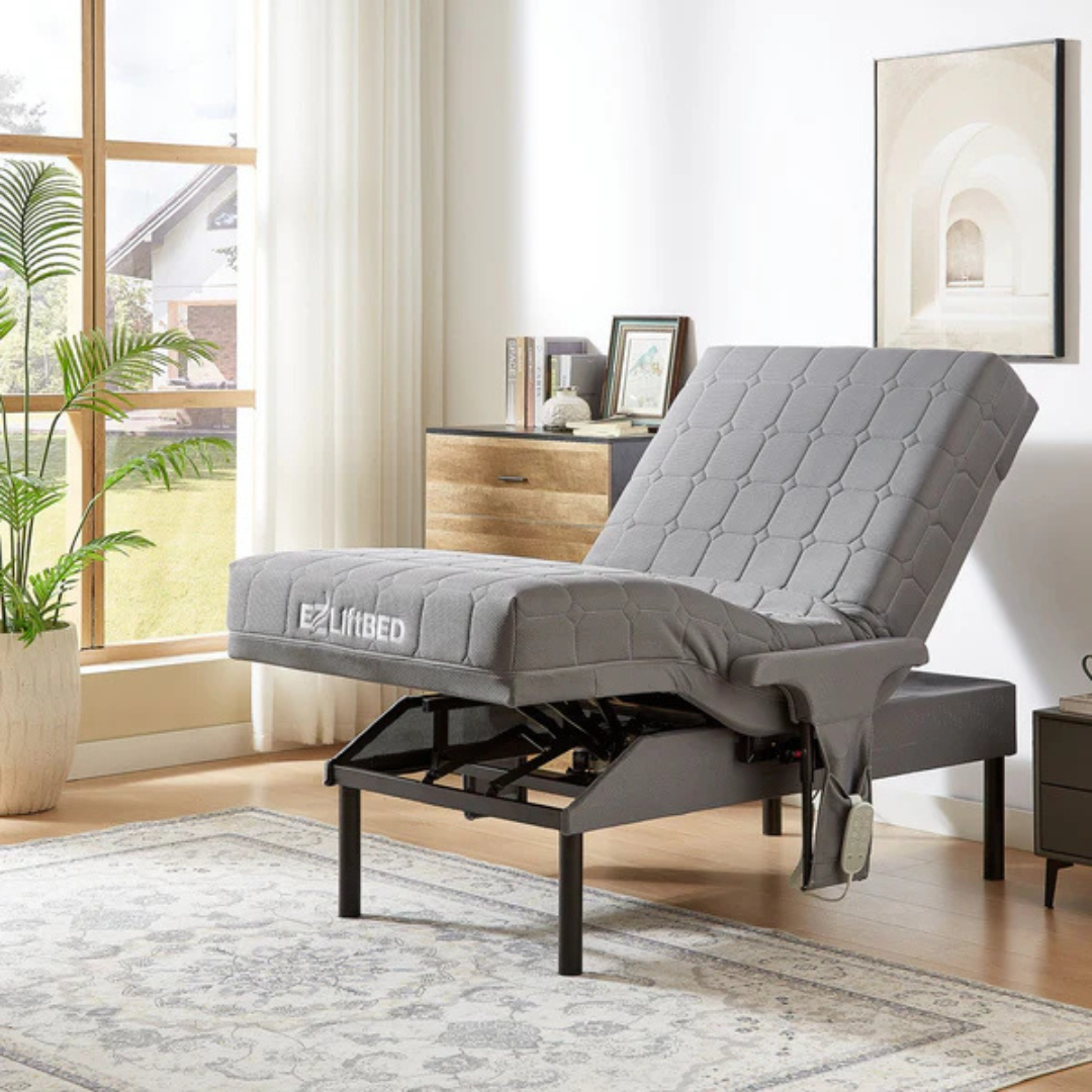 The EZLift Bed™ - Deluxe Sleep To Stand Electric Bed with a gray quilted mattress is elevated at the head and foot in a bright room featuring large windows, a patterned rug, dresser, plants, and decorative wall art.