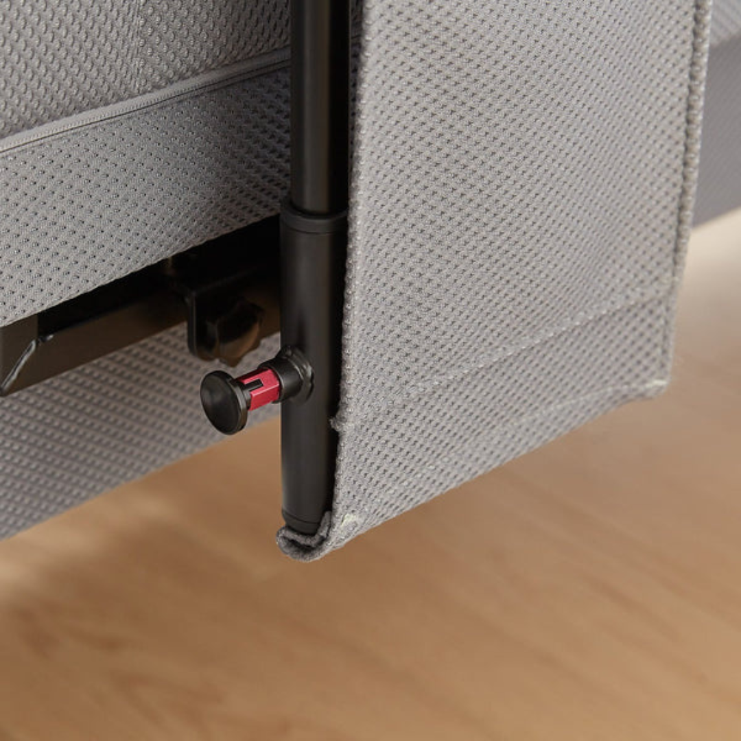 A close-up of the EZLift Bed™ - Deluxe Sleep To Stand Electric Bed’s metal support leg with a red lock, partly hidden by a gray dust cover, on a wooden floor.