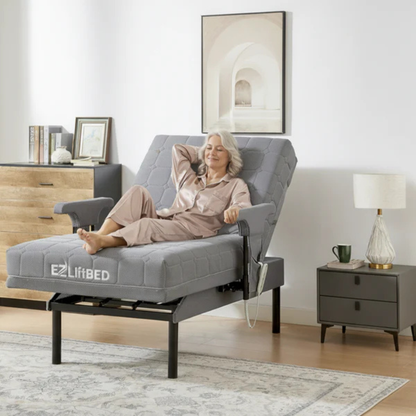 An older woman sits comfortably on an EZLift Bed™ - Deluxe Sleep To Stand Electric Bed in a modern bedroom, smiling and holding a remote. The room features light-colored walls, wooden furniture, and minimalistic decor.