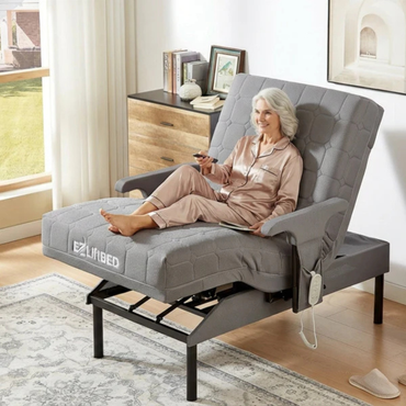 An older woman in satin pajamas smiles as she relaxes on a gray EZLift Bed™ - Deluxe Sleep To Stand Electric Bed, using the remote. The bed is elevated at the head and legs in a bright, cozy room with a rug, dresser, and decor.