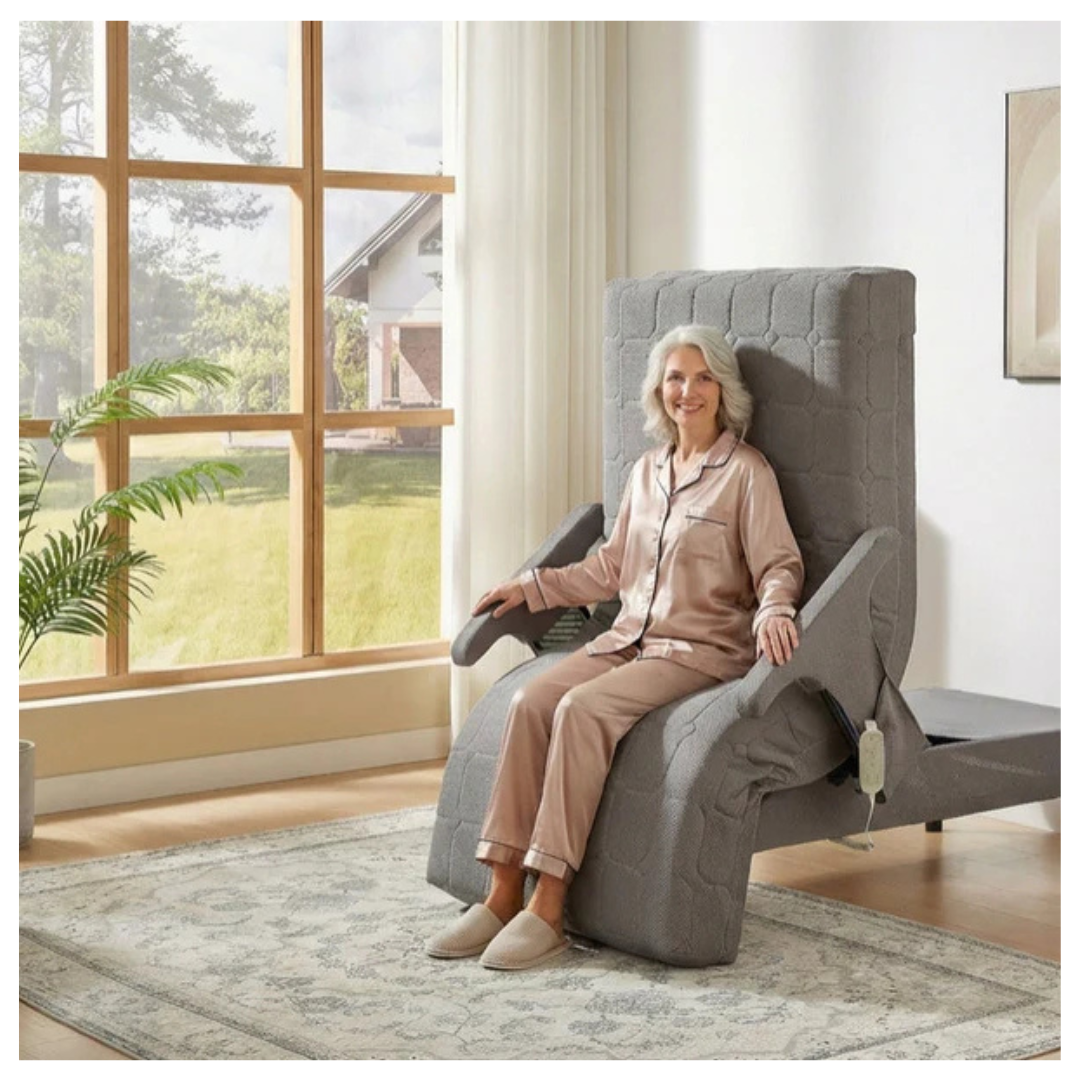 An older woman in pajamas and slippers relaxes on a gray recliner next to the EZLift Bed™ - Deluxe Sleep To Stand Electric Bed in a bright living room with a large window and light-colored rug.