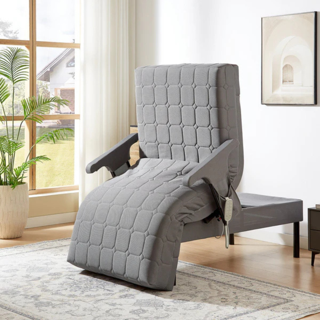 A gray, quilted motorized reclining chair sits in a bright living room with a large window, plant, and art. Inspired by the EZLift Bed™ - Deluxe Sleep To Stand Electric Bed, it features a handheld remote on the arm for simple control.