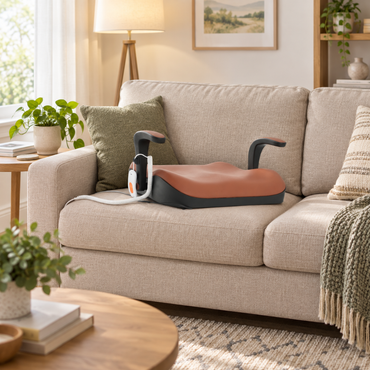 A brown and black booster seat with armrests sits on a beige couch next to the Easy Stand-Up Electric Lift | VOCIC AX16 Pro recliner in a cozy living room featuring plants, a lamp, a throw blanket, and framed wall art.