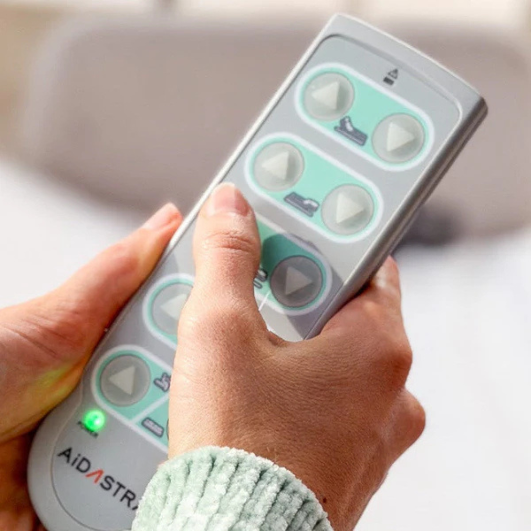 A person in a textured light green sleeve holds a remote labeled AIDASTRA, featuring arrow buttons and a green indicator light, for the Elevra Deluxe Rotating Hi-Lo Lift Assist Bed.