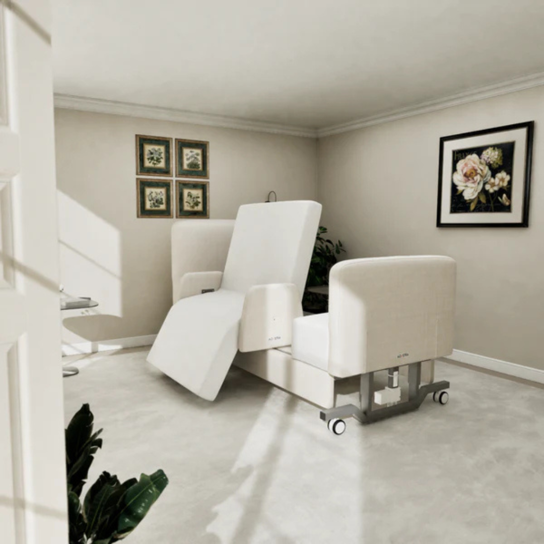 A modern beige reclining chair with wheels sits in a bright, minimalist room with light walls, floral art, framed photos, and plants—perfectly complementing the Elevra Deluxe Rotating Hi-Lo Lift Assist Bed's comfort.