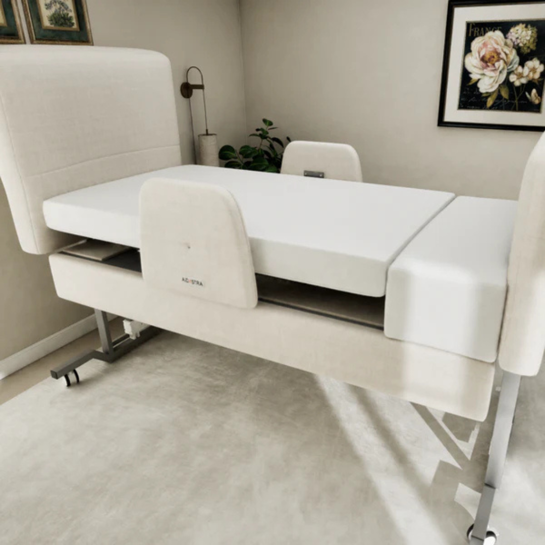 The Elevra Deluxe Rotating Hi-Lo Lift Assist Bed, featuring beige upholstery and split head and footrests, is set in a carpeted room accented by framed floral art and a potted plant.