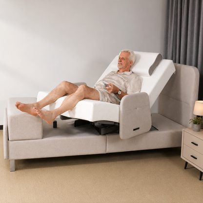 An elderly man in light pajamas reclines on the Elevra Deluxe Rotating Lift Assist Bed - Dual King, using a remote to tilt his head and legs. A nightstand with a lamp and potted plant sits beside this advanced homecare bed.