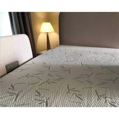 A close-up of a bed with the Waterproof Mattress Shield For Elevra Beds - PureBamboo™, showcasing its light color and subtle leaf pattern next to a beige headboard, with a lamp and curtain softly lit in the background.