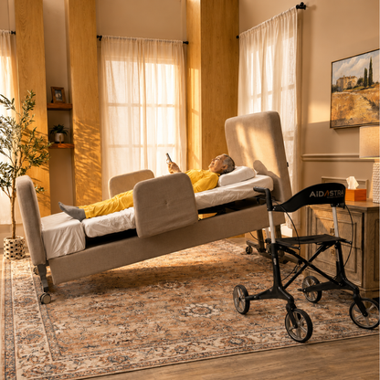 An elderly person in yellow pajamas lies in a beige Elevra Deluxe Rotating Hi-Lo Lift Assist Bed, using a smartphone. The bed is tilted, with a black wheelchair nearby in a sunlit room with large windows and warm decor.