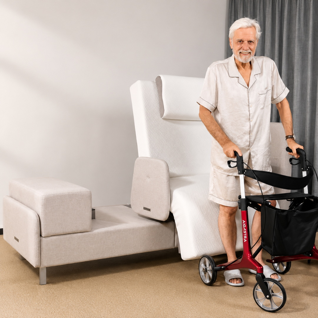Smiling elderly man in pajamas stands with a red walker beside an Elevra Deluxe Rotating Lift Assist Bed - Dual King and a small upholstered bench in a bright, carpeted room.