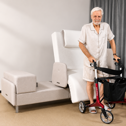 Smiling elderly man in pajamas stands with a red walker beside an Elevra Deluxe Rotating Lift Assist Bed - Dual King and a small upholstered bench in a bright, carpeted room.