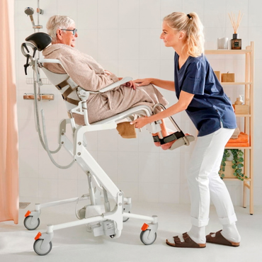 A caregiver adjusts the footrests of an Etac Alto Electrically Powered Shower Commode Chair occupied by a person in a bathrobe.