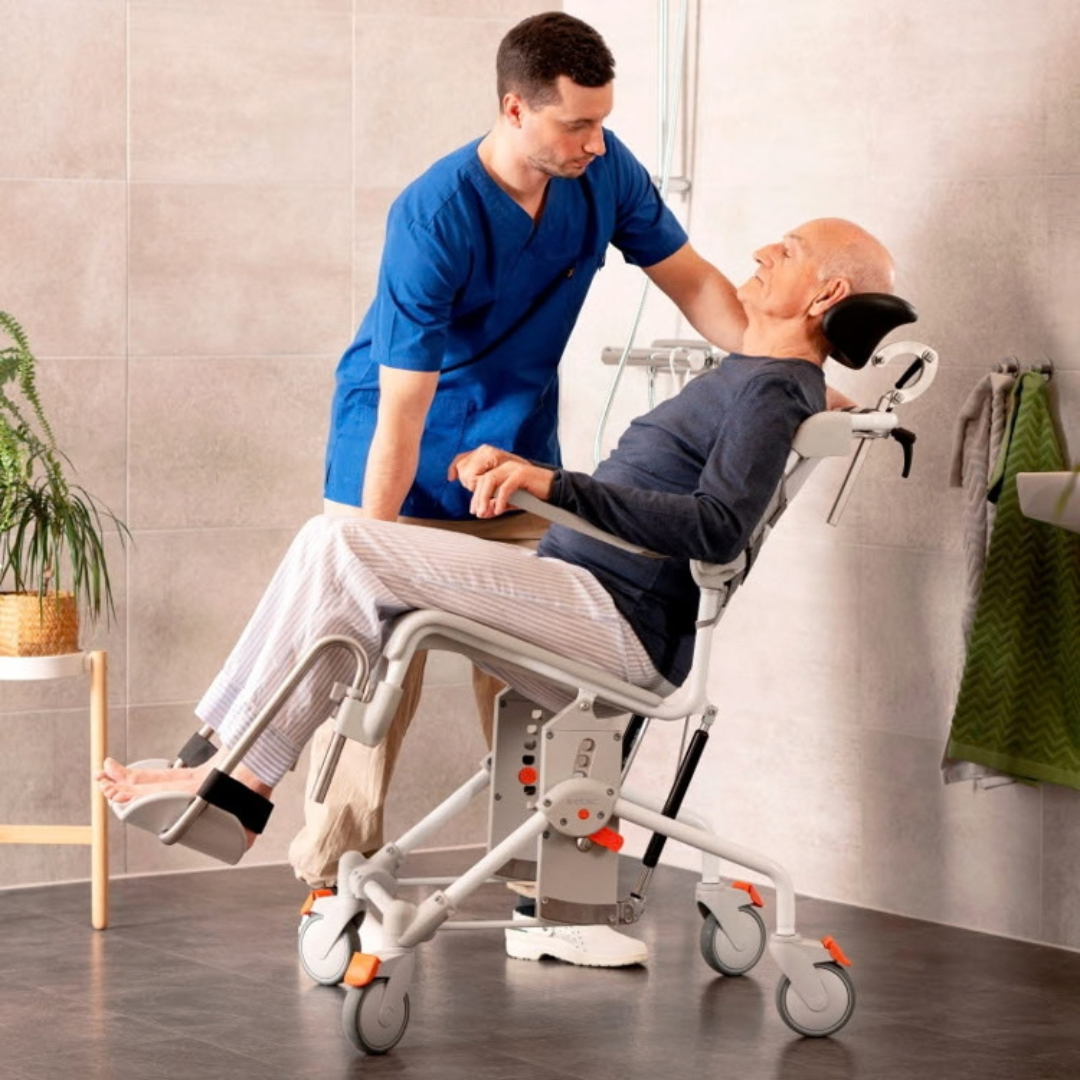 A caregiver assists an elderly man seated in an Etac Swift Mobil Tilt-2 Tilt-In-Space Mobile Shower Commode.