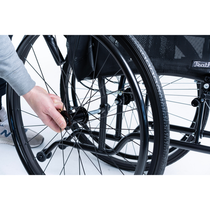 A person’s hand grasps the wheel of a Feather HD Ultralight Bariatric Wheelchair (22 lbs, black, 24-inch), adjusting or detaching it. The sturdy frame and spokes stand out against a white background.