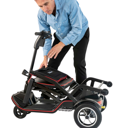 A person in a blue shirt bends over to fold the Feather Ultralite Portable Scooter—this black and red, three-wheeled mobility scooter weighs only 37 lbs and sits on a white background.