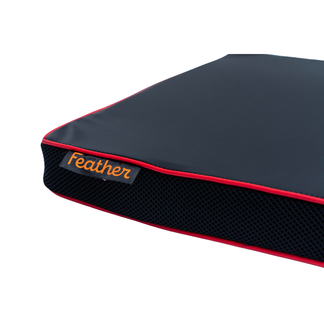 A close-up of the Feather Portable Cushions—an all-purpose black wheelchair cushion with red piping, mesh sides, water-resistant fabric, and a handle. An orange tag reads "Feather." Photographed on a white background.