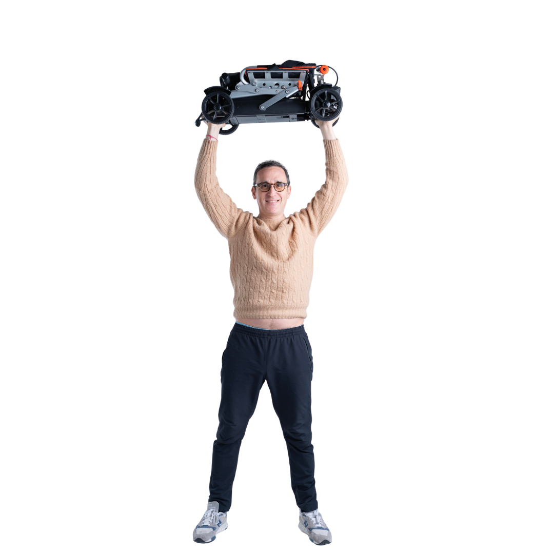 A smiling person in glasses, a beige sweater, dark pants, and sneakers holds the Feather 27X Folding Portable Mobility Scooter—only 27 lbs—above their head against a white background.
