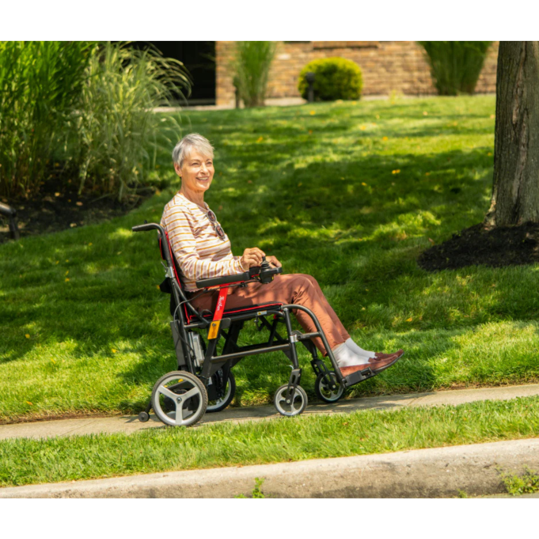 An older woman with short gray hair smiles while sitting in the Feather Powerchair—an ultralight, travel-approved electric wheelchair weighing only 33 lbs—on a sunny sidewalk surrounded by grass and trees.