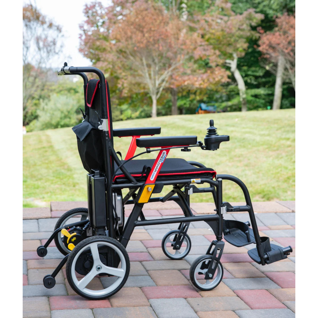 The Feather Powerchair - Ultralight Travel Approved Electric Wheelchair (only 33 lbs) in black and red, with padded seat, armrests, controls, and footrests visible on the right side, is positioned on a patio with grass and trees in the background.