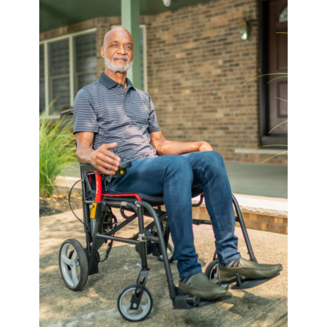 An older man in a Feather Powerchair – Ultralight Travel Approved Electric Wheelchair (only 33 lbs) smiles outside a brick house. The lightweight powerchair blends seamlessly with the greenery and porch behind him.