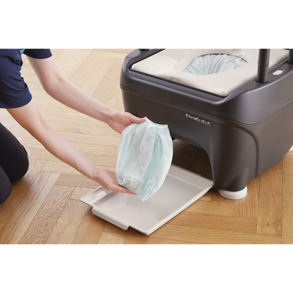 A person kneels on a wooden floor, removing a sealed waste bag from a gray portable toilet, demonstrating the sanitary sealing and easy replacement of the Film Cassette for Wrappon ORB Commode.