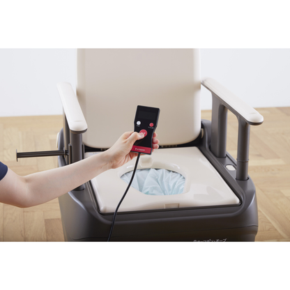 A person holds a remote beside a beige and brown commode with armrests and an open seat, lined with the Film Cassette for Wrappon ORB Commode, providing a modern portable toilet solution on a wooden floor.