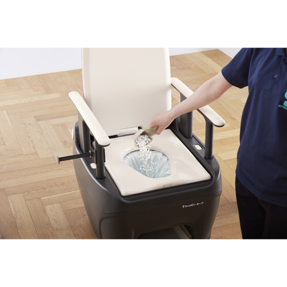 A person pours white powder into the bowl of a portable commode with armrests, likely as part of using the Film Cassette for Wrappon ORB Commode, on a wooden floor.