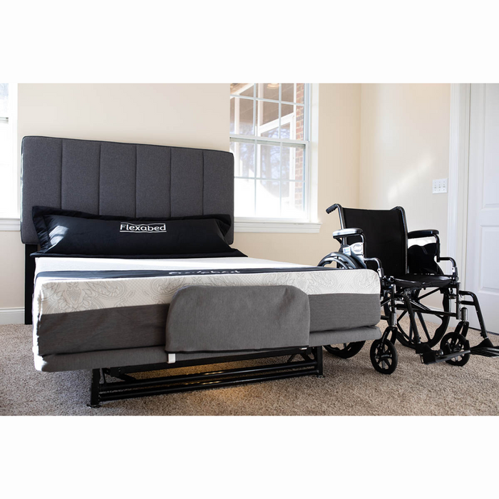 The Flexabed Hi-Low SL Adjustable Bed, a luxury hospital bed alternative with a modern gray and white design, sits next to a black wheelchair in a bright room. Sunlight illuminates the dark gray headboard, Flexabed pillow, and carpeted floor.