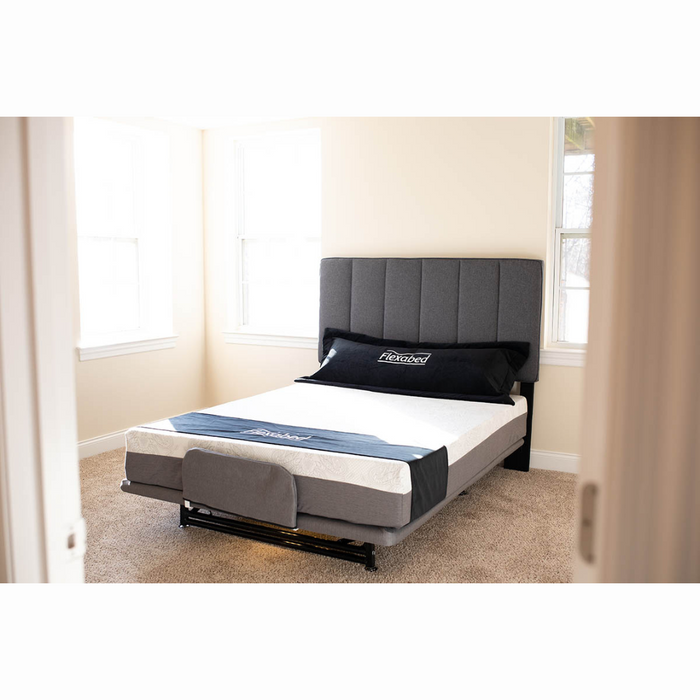 In a bright room with light walls and a beige carpet, the Flexabed Hi-Low SL Adjustable Bed features customizable height settings. The dark gray upholstered headboard and base complement its white mattress with back support, topped with a neatly folded blue blanket at the foot.