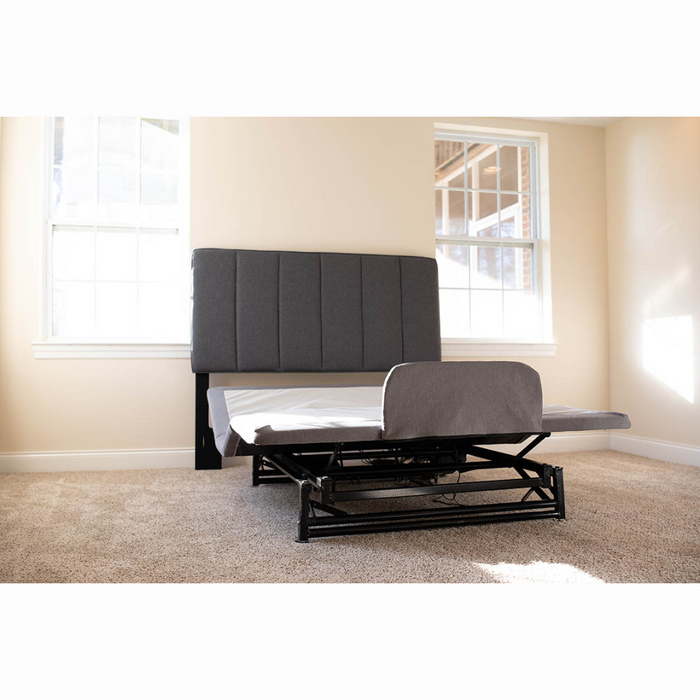 The Flexabed Hi-Low SL Adjustable Bed, a luxury alternative to a hospital bed, sits partially elevated on light brown carpet in a sunlit room. Its grey upholstered headboard against the cream wall with large windows offers ultimate comfort and customizable back support.