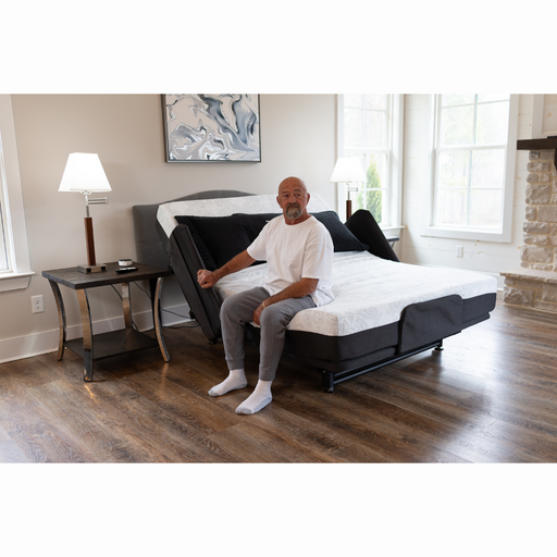 A man sits on a Flexabed Hi-Low SL Adjustable Bed, luxurys alternative to a hospital bed, with the backrest raised in his brightly lit bedroom. Dressed in a white shirt and gray pants, he enjoys custom back support beside a lamp-topped nightstand and wall-hung painting.