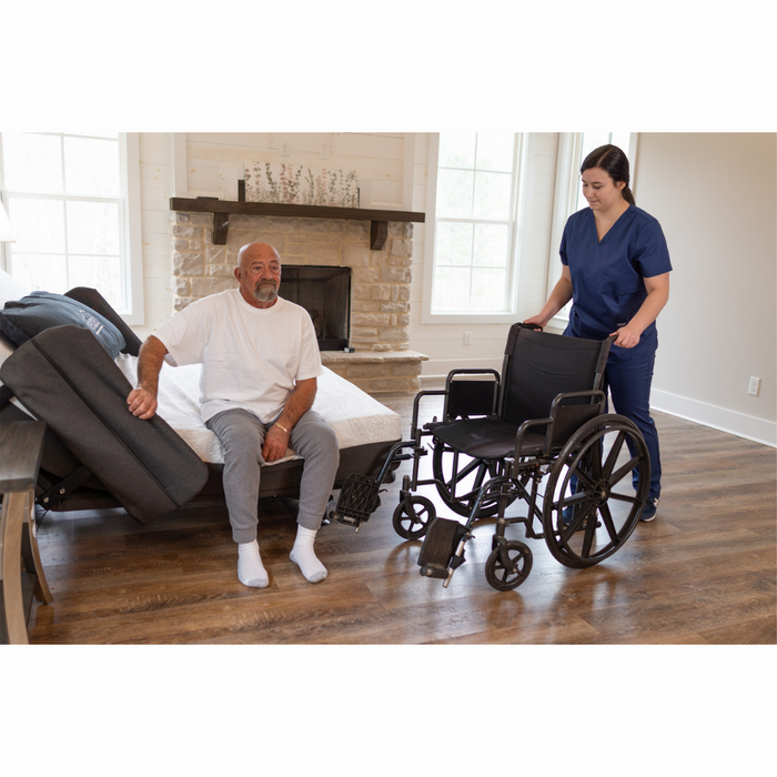 A woman in blue scrubs helps an elderly man from a Flexabed Hi-Low SL Adjustable Bed to a wheelchair in a cozy living room with hardwood floors and a fireplace. The man, in a white shirt and gray pants, enjoys the customized back support as sunlight floods the room.