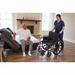 A woman in blue scrubs helps an elderly man from a Flexabed Hi-Low SL Adjustable Bed to a wheelchair in a cozy living room with hardwood floors and a fireplace. The man, in a white shirt and gray pants, enjoys the customized back support as sunlight floods the room.