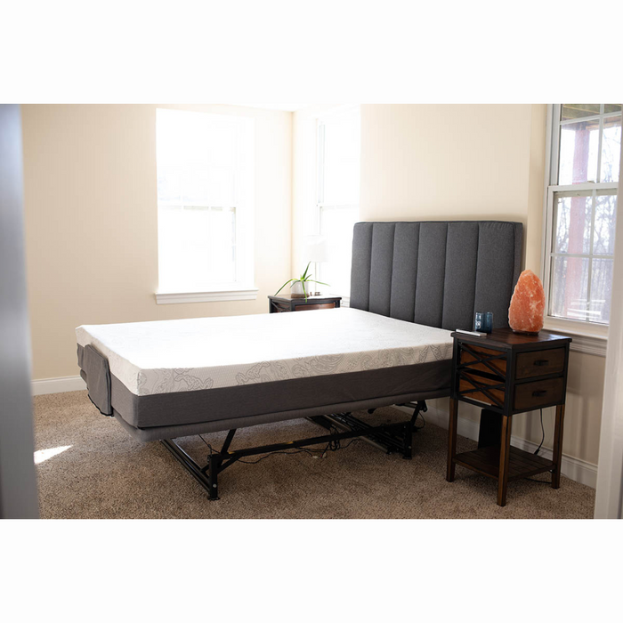 A modern bedroom features a Flexabed Hi-Low SL Adjustable Bed with customizable back support and a white mattress, positioned by two large windows. A bedside table holds a lamp, small plant, and salt lamp on the light brown carpet completing the serene setting.