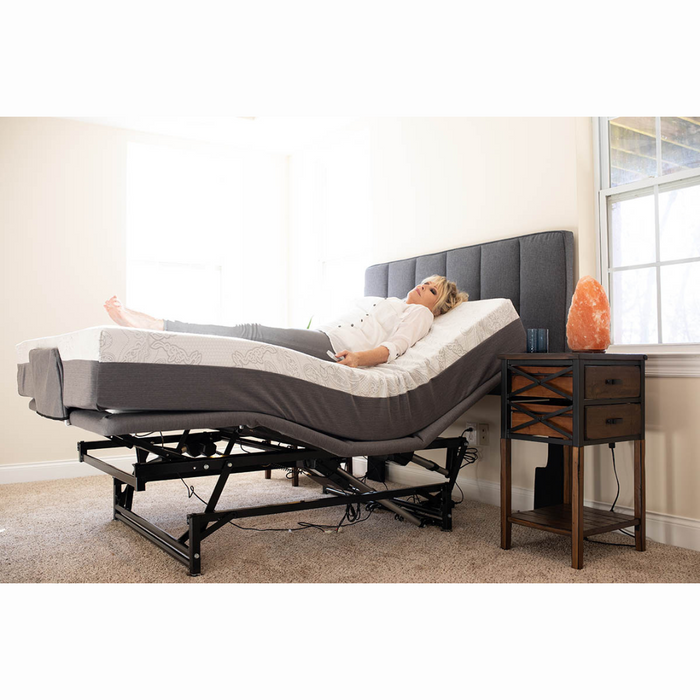 A woman lies on a Flexabed Hi-Low SL Adjustable Bed, head raised for customized back support. Shes in a bright room with beige walls and a window. A wooden bedside table holds a Himalayan salt lamp next to the bed, revealing its adjustable base beneath.