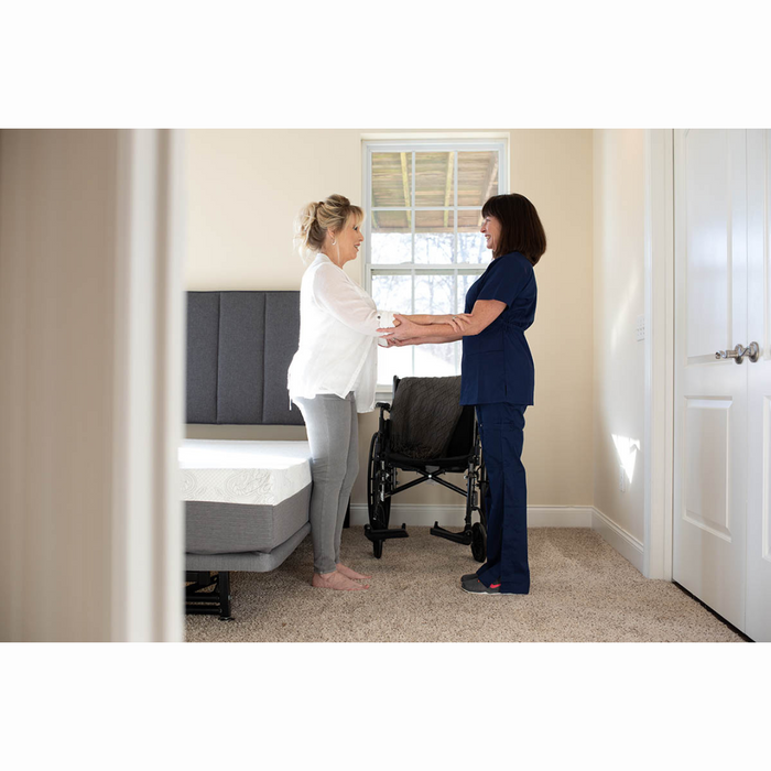 In a sunlit bedroom, a woman dressed in a white blouse and gray pants stands near the Flexabed Hi-Low SL, offering customized back support. She is assisted by a healthcare worker in blue scrubs, with an adjustable-height bed and wheelchair nearby, creating a warm atmosphere.