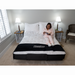 A woman in pink pajamas relaxes on her Flexabed Premier Adjustable Full Electric Bed with Massage, reading a book. The bed, dressed in white bedding and pillows, is flanked by two bedside tables with lamps and a chair, creating a cozy bedroom ambiance.