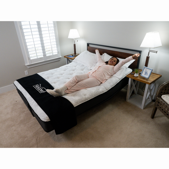 A person stretches in a bright room on a Flexabed Premier Adjustable Full Electric Bed with Massage, featuring dual-timed massage settings and topped with a textured white cover and dark blanket. A remote is nearby, while twin bedside tables hold lamps and decorations.