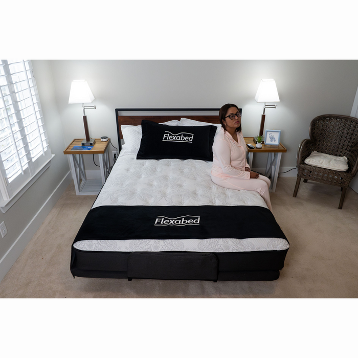 A person in pajamas sits on the edge of a Flexabed Premier Adjustable Full Electric Bed with Massage in a softly lit bedroom. The room includes customizable configurations like a nightstand with a lamp and a decorative chair, while the bedding displays Flexabed branding.