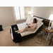 A woman in pink pajamas reads a book while reclining on the Flexabed Premier Adjustable Full Electric Bed with Massage, enjoying its customizable settings. The room features beige carpet, a nightstand with a lamp and photo frame, plus a window with shutters. A black foot cover and slippers rest on the floor.