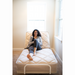 In a bright room with large windows, a curly-haired person relaxes on a Flexabed Value-Flex Adjustable Bed. Casually dressed in a gray hoodie and leggings, they sit comfortably, smiling and holding a mug while enjoying the adjustable height and relaxed recline features of their bed.