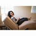 A smiling woman with curly hair relaxes on a Flexabed Value-Flex Adjustable Bed, holding a remote. She wears casual clothes in a softly lit room with natural light streaming through large windows in the background.