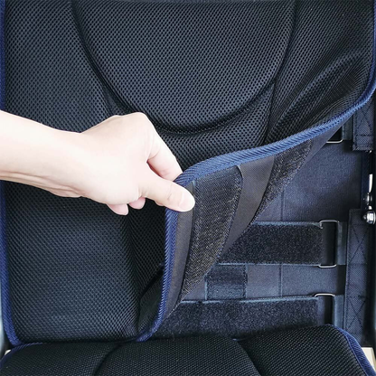 A hand lifts the edge of a black, padded seat cover with velcro fastenings on the Foldawheel ECO Lightweight Folding Powerchair, revealing the seat structure and straps underneath.