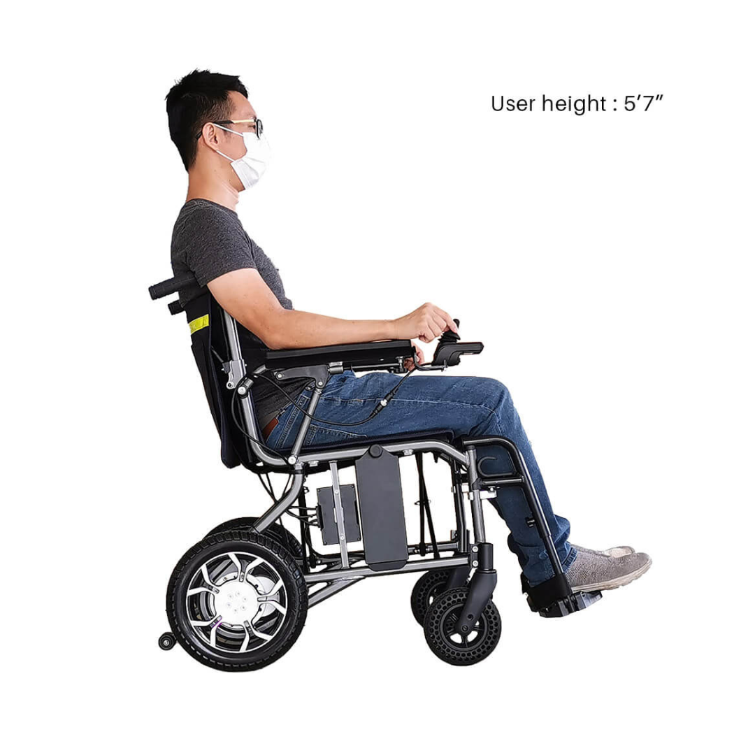 A man in a face mask and glasses, wearing a gray t-shirt and jeans, sits in a Foldawheel ECO Lightweight Folding Powerchair. Text reads "User height: 5'7"." He is shown in profile against a white background.