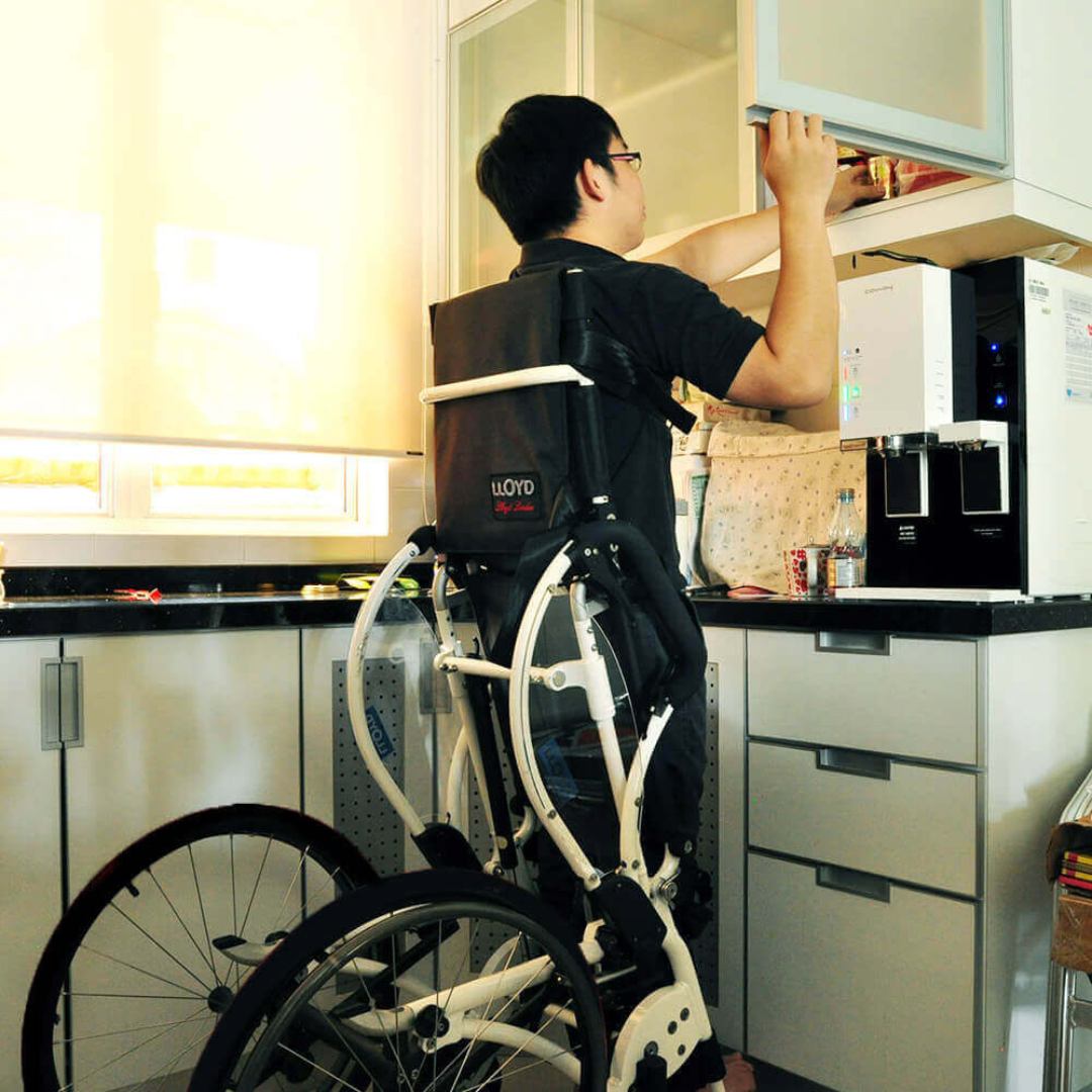 A person using the Foldawheel Lloyd LEO Ultralite Standing Manual Wheelchair reaches into an upper kitchen cabinet, showing how this lightweight chair increases accessibility in the kitchen.