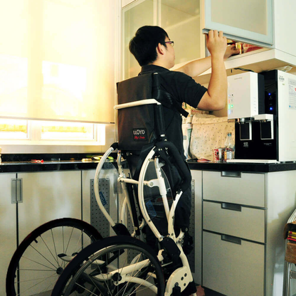A person using the Foldawheel Lloyd LEO Ultralite Standing Manual Wheelchair reaches into an upper kitchen cabinet, showing how this lightweight chair increases accessibility in the kitchen.