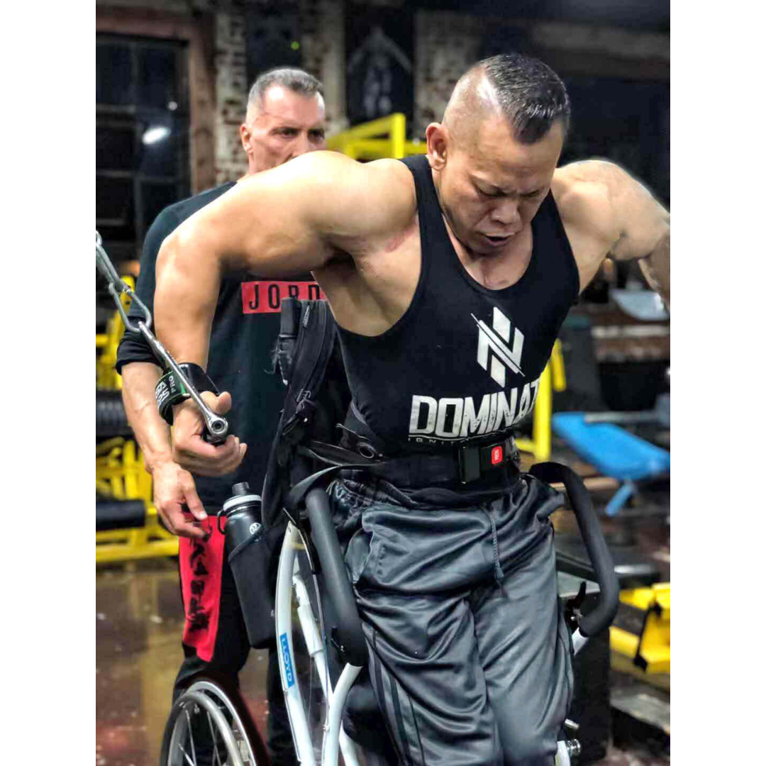 A muscular man in a Foldawheel Lloyd LEO Ultralite Standing Manual Wheelchair works out on a cable machine wearing a black “Dominate” tank top, while another man assists him in the gym.
