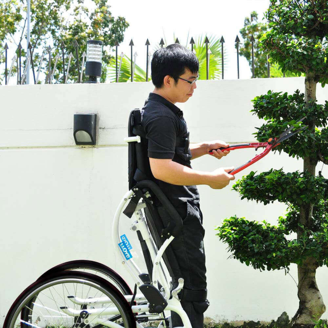 Using a Foldawheel Lloyd LEO Ultralite Standing Manual Wheelchair, a man trims a neatly shaped bush in a garden, reaching easily thanks to the standing feature; white wall and green trees appear in the background.