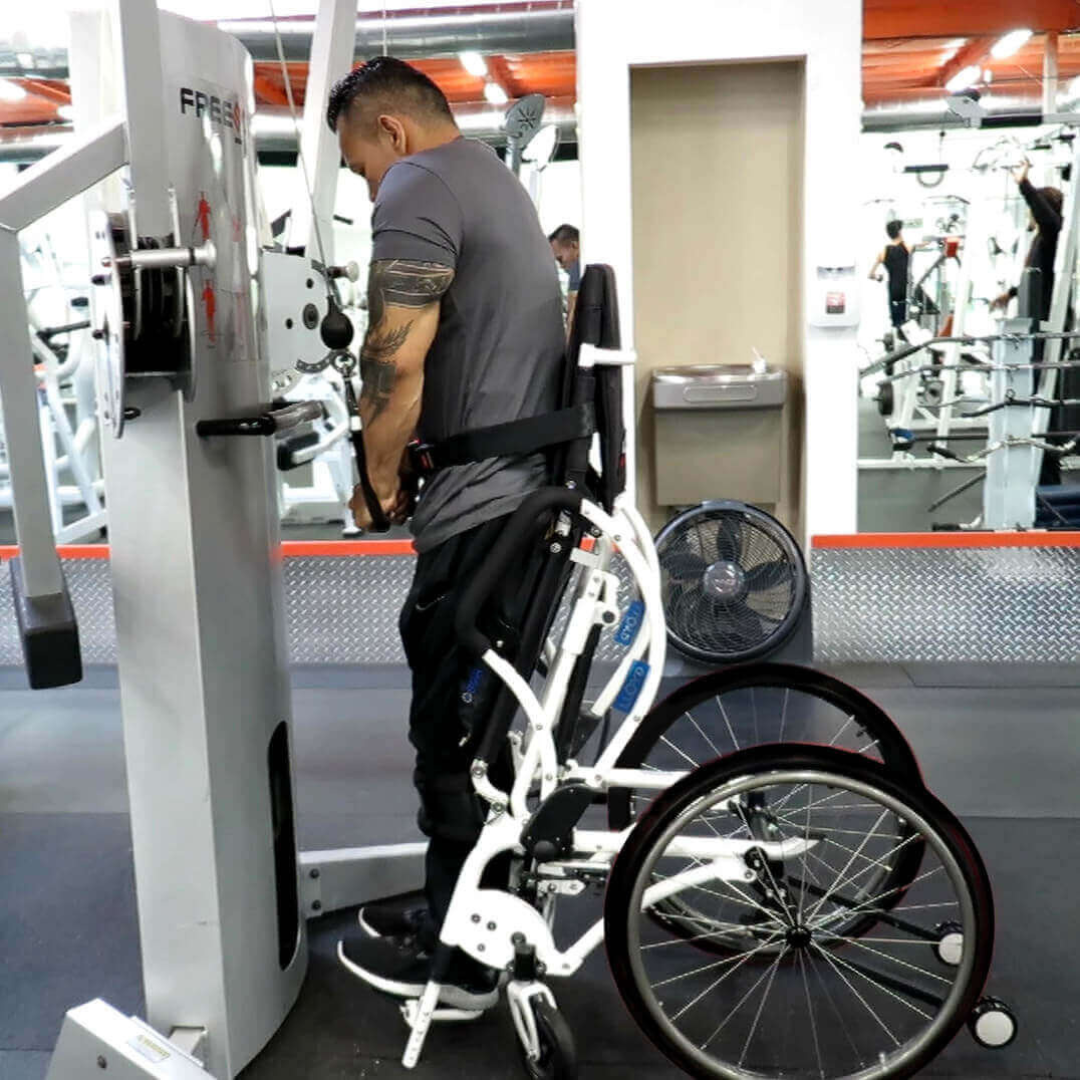 A man uses the Foldawheel Lloyd LEO Ultralite Standing Manual Wheelchair to exercise with gym equipment, gripping the handles and facing a machine, with workout machines and mirrors visible in the background.
