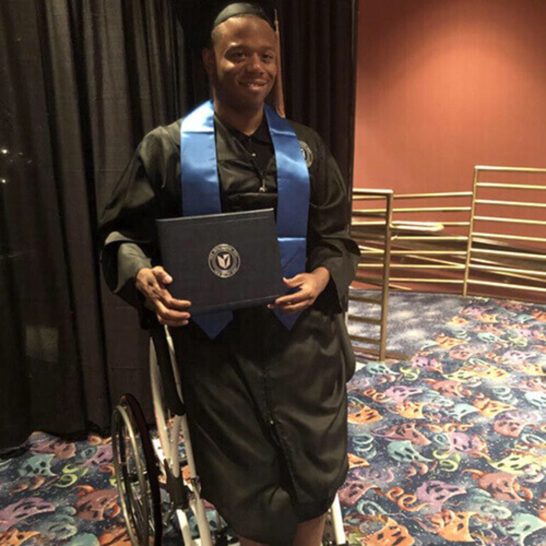 A smiling graduate in a cap and gown sits in a Foldawheel Lloyd LEO Ultralite Standing Manual Wheelchair, holding a diploma folder. He wears a blue stole, with colorful carpet and theater-style railings behind him.